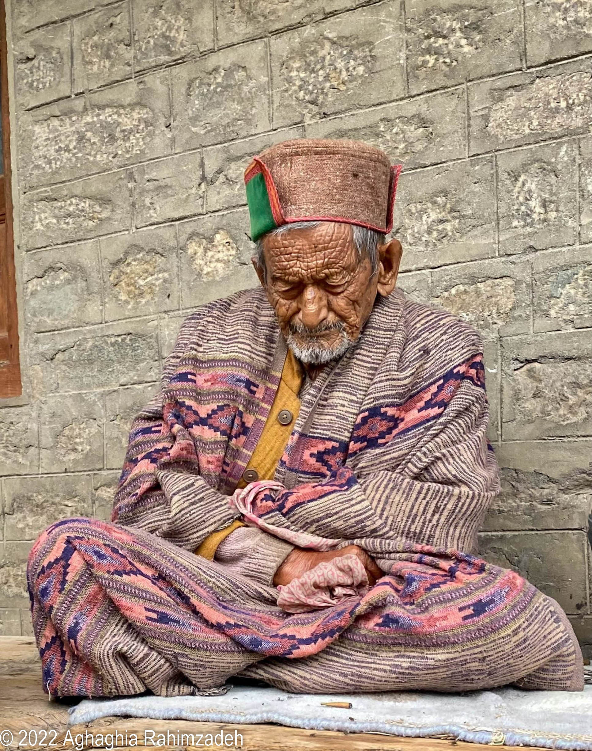 An elderly man sits cross-legged wearing a colorful woven robe and hat. 